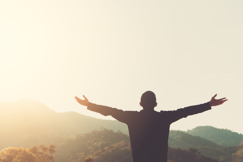 Silhouette of a man raising his hands up to the sky at sunset, foothills visible in the background.