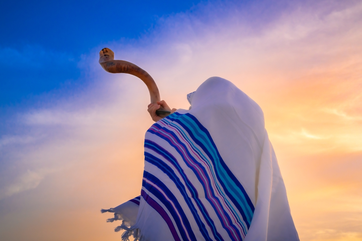 Person covered in a tallit blowing a shofar at sunset.